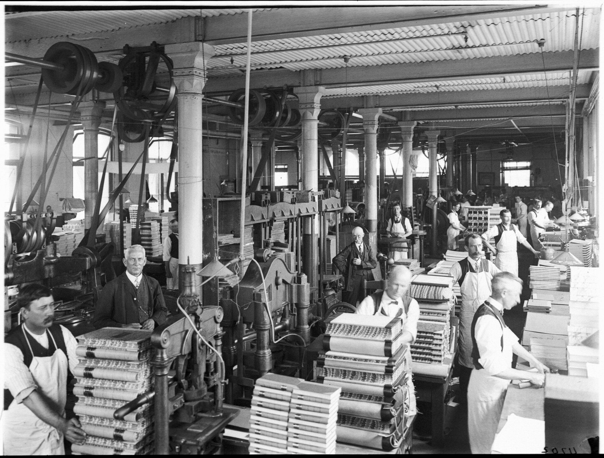 Men in work aprons stand in a workshop behind stacks of large hardcover books