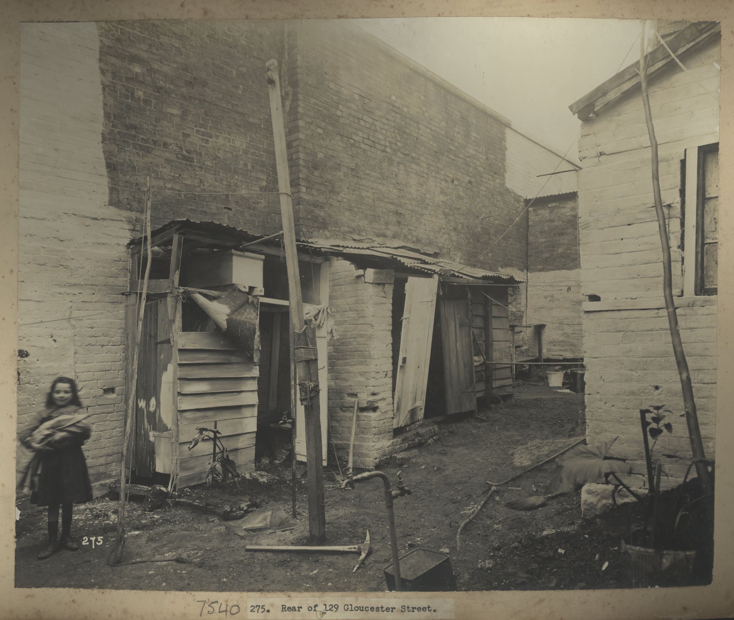 A young girl stands to the left of a backyard holding a baby. Wooden extensions from the brick building are in disrepair and the doors are falling off