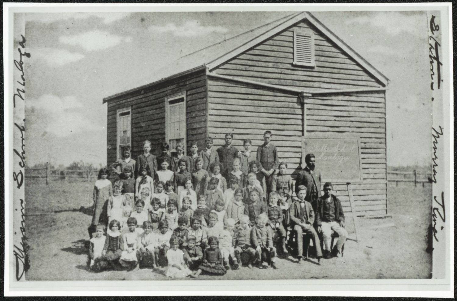 An outdoor class photo of students and their teacher in front of a small wooden school building