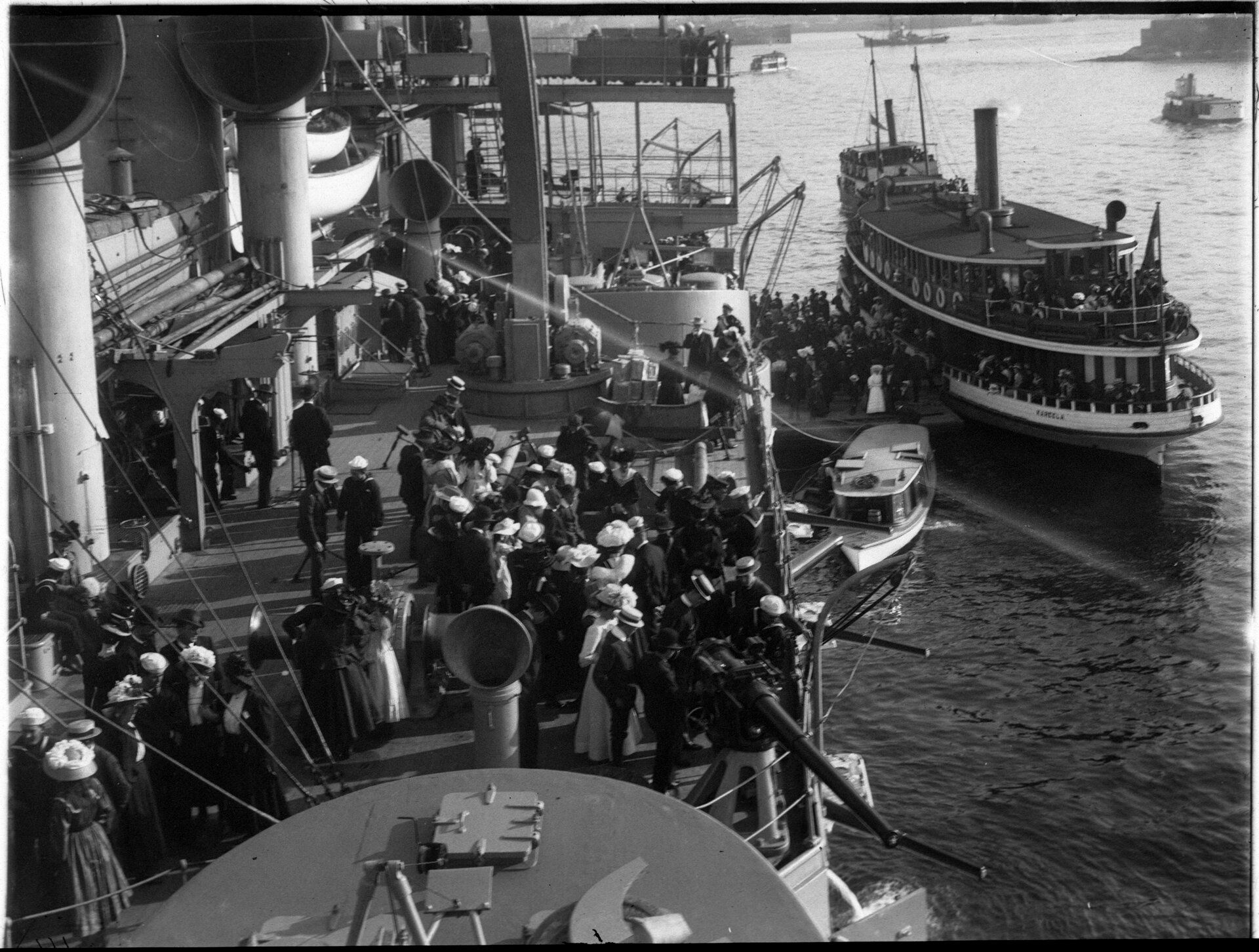  Crowds of visitors fill the deck of a ship. More visitors arrive from a launch vessel in the background