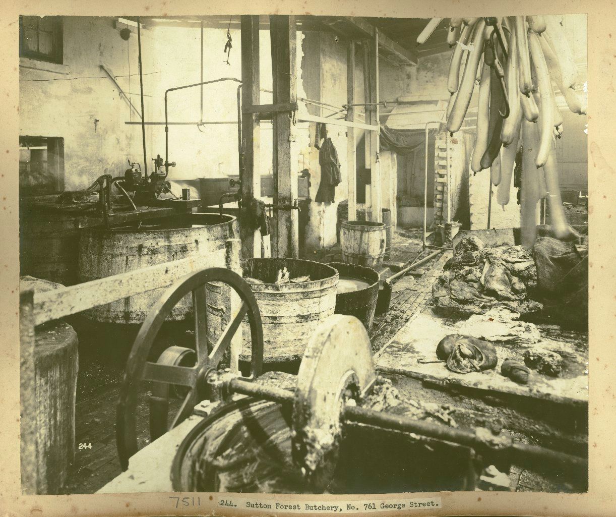 A kitchen in a butchery showing wooden barrels of liquid and sausages hanging above a wooden work bench
