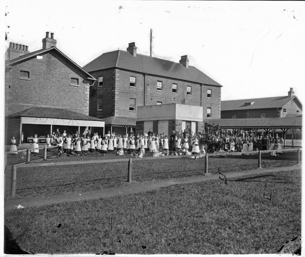 School girls in white smocks stand in a large group outside a brick building