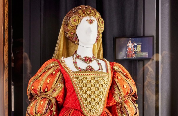 A mannequin dressed in an elaborate Renaissance-style stage costume stands in a large Perspex-fronted display case . On the rear wall a small screen is showing a scene from the opera Lucrezia Borgia where Dame Joan Sutherland is wearing the same costume.