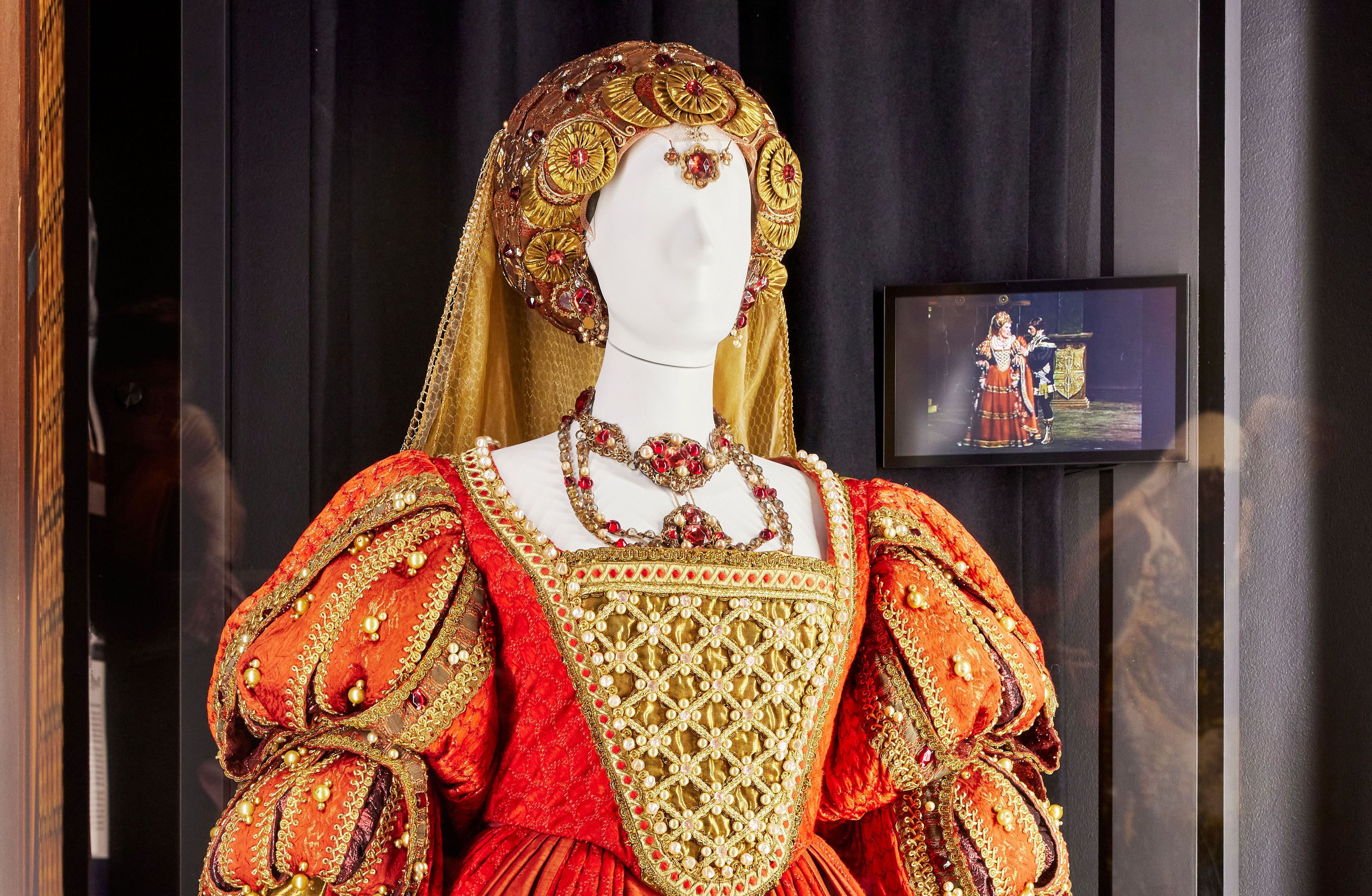 A mannequin dressed in an elaborate Renaissance-style stage costume stands in a large Perspex-fronted display case . On the rear wall a small screen is showing a scene from the opera Lucrezia Borgia where Dame Joan Sutherland is wearing the same costume. 