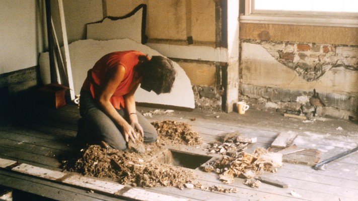 Person in red shirt kneeling in front of lifted floorboards removing debris.