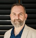 Man in a blue shirt and tan sports coat with a beard.