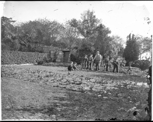 A man holding a garden hoe stands with a group of boys who are learning gardening