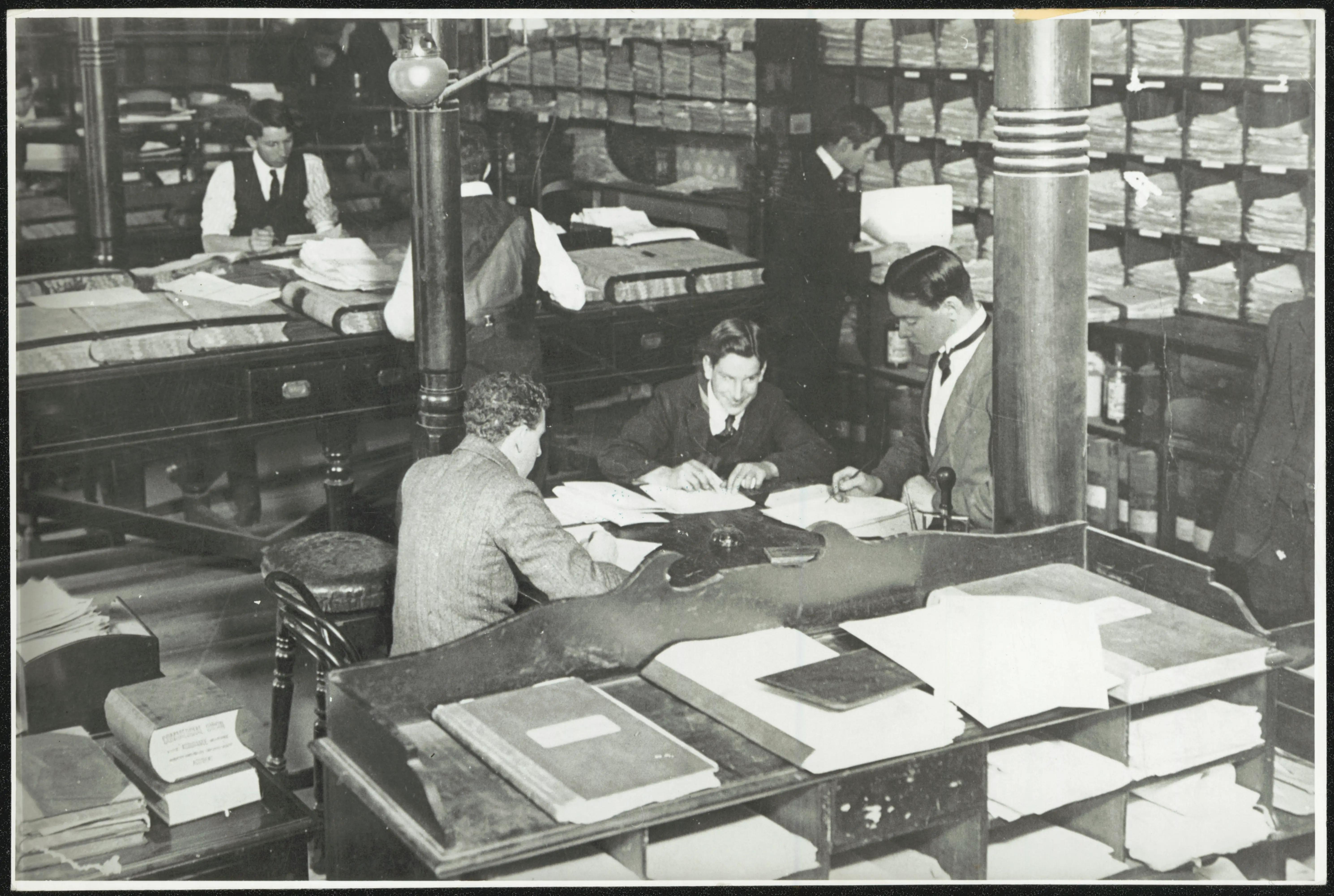 Black and white photo of clerks working at desks in an office, surrounded by large ledgers and pigeon holes and paperwork