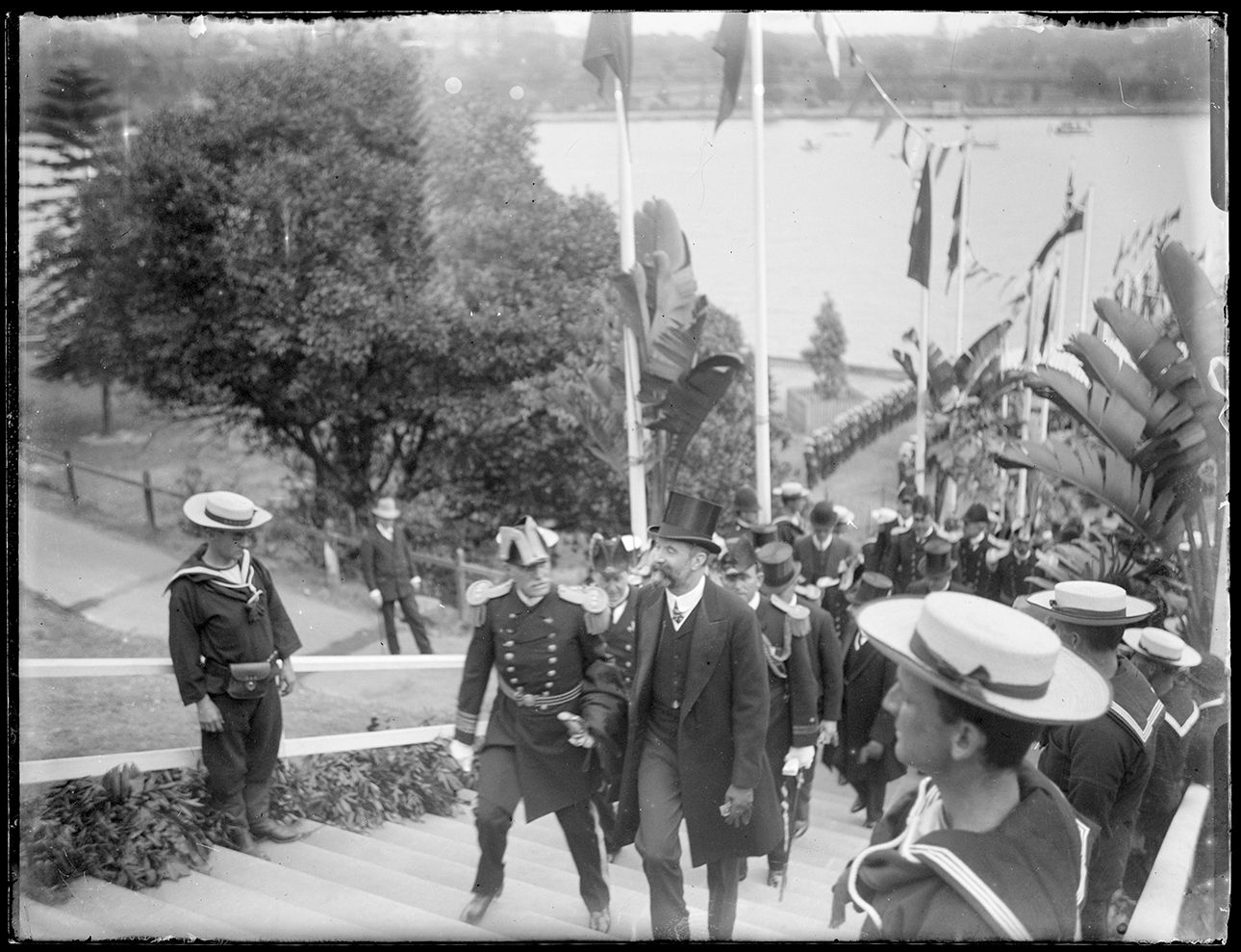 Sailors in boater hats line either side of stairs leading to a harbour and a group of men in top hat and tails walk up with stairs with an admiral in full regalia 