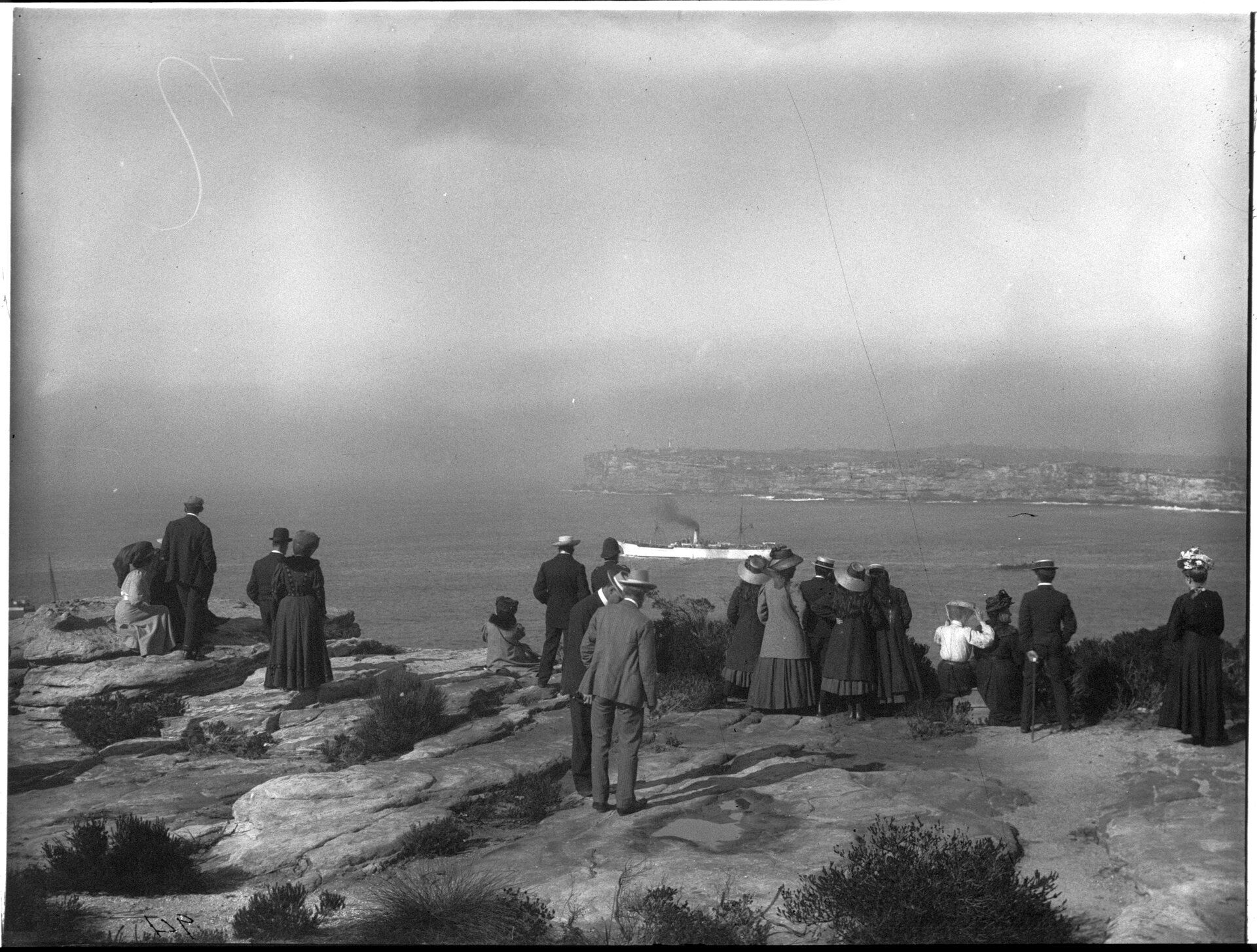  A crowd stand on a harbour clifftop watching a ship depart