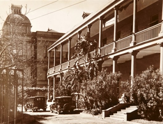 Large 2 storey building with deep verandahs, steps leading to lower verandah and bushes and driveway in the foreground.
