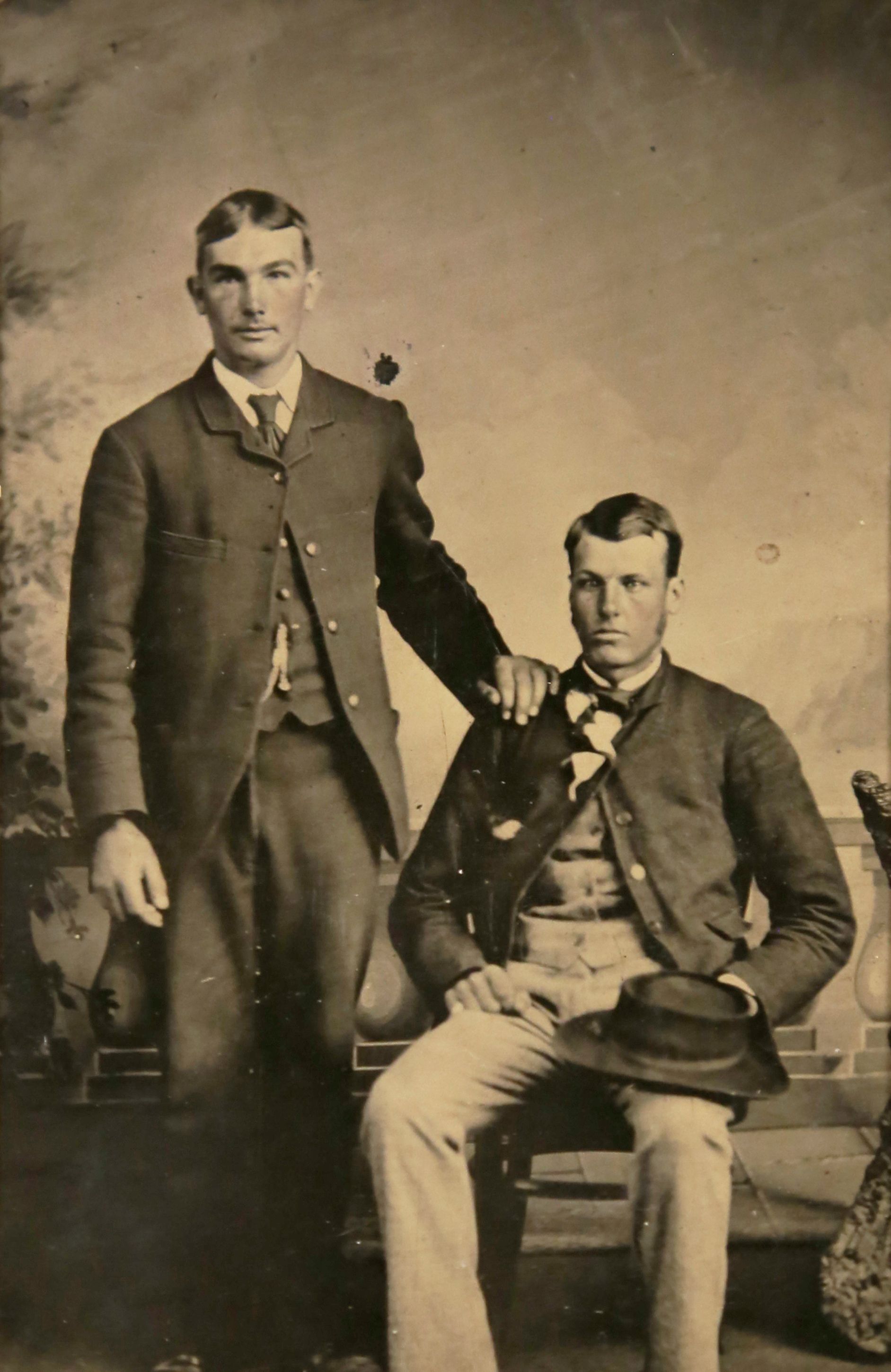 Portrait of two men. One sits holding his hat on his leg and the other other stands, resting his hand on the shoulder of the seated man