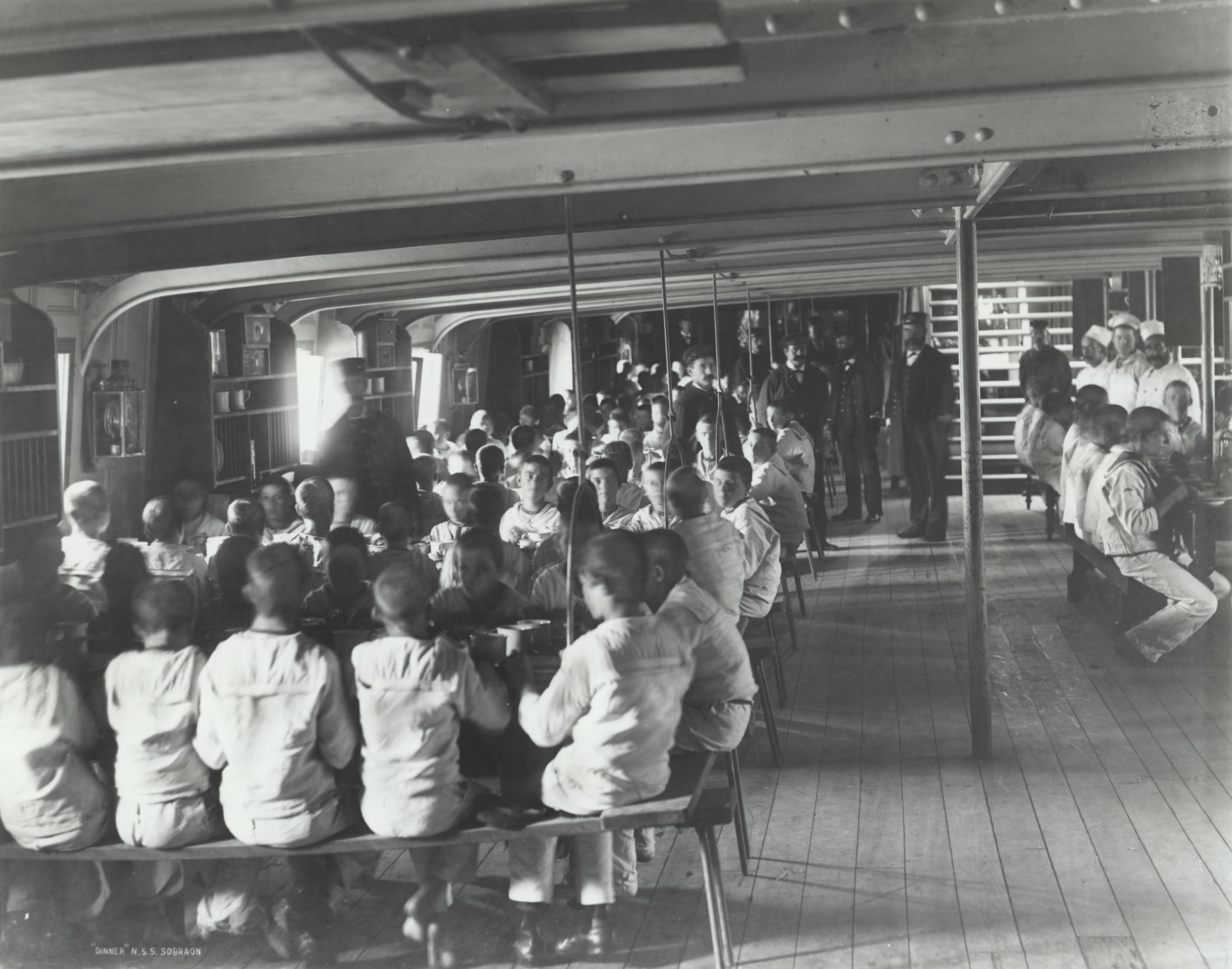 Boys in nautical clothing sit at long tables in a ship's lower deck  