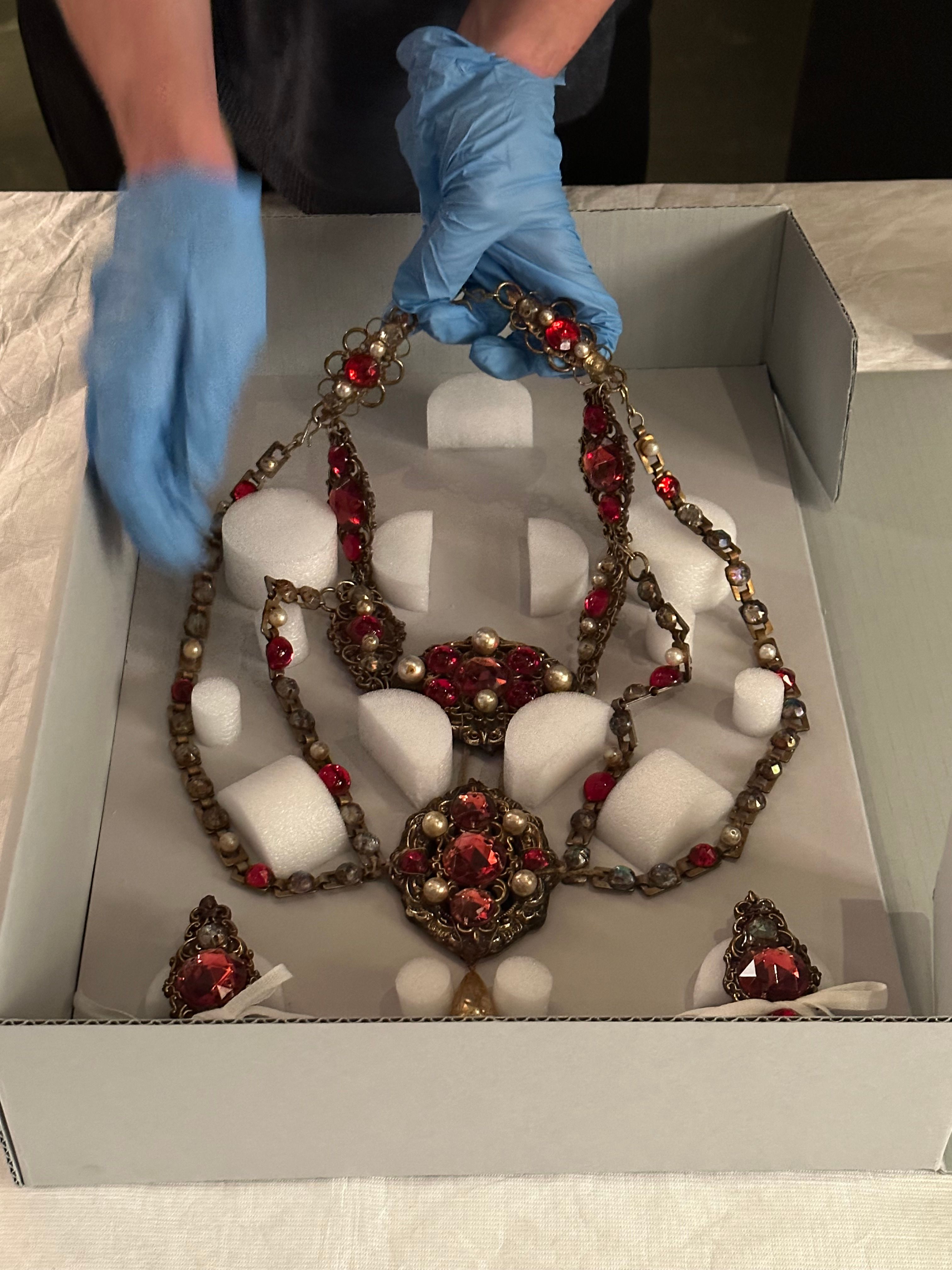 An elaborate multi-strand costume necklace made to resemble gold, pearls and rubies is being removed from a grey cardboard box. The box has many small shaped segments of foam set within it which hold the necklace segments in place.   