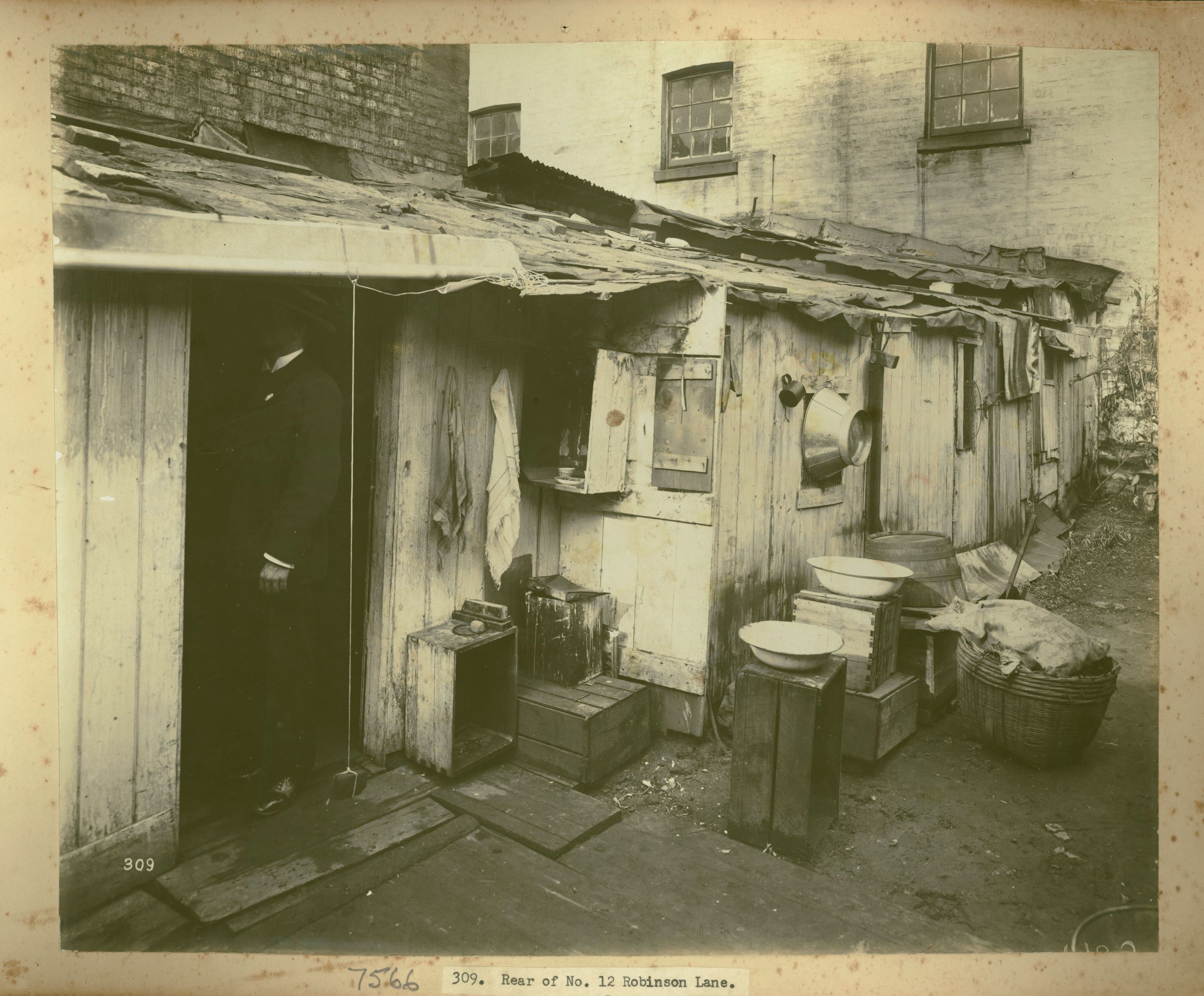A man stands in the doorway at the rear of a house. Outside are pots and large bowls sitting on upturned wooden crates