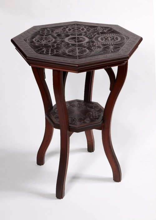  Chip-carved table  A demountable, chip-carved occasional table of kauri pine, made by carpenter George Stevenson Liggins (1874 -1907), Melbourne, c1902. 80(h) x 59(w) x 59cm(d).  Timbers chosen for chip-carving in Australia were typically softwoods such as pine, maple or cedar. In this example, the table tops are constructed from Kauri Pine, along with the two larger supports, while the two smaller supports (not visible in this photo) are constructed from Queensland Maple and were probably added at a later date to stem warping of the larger top.  