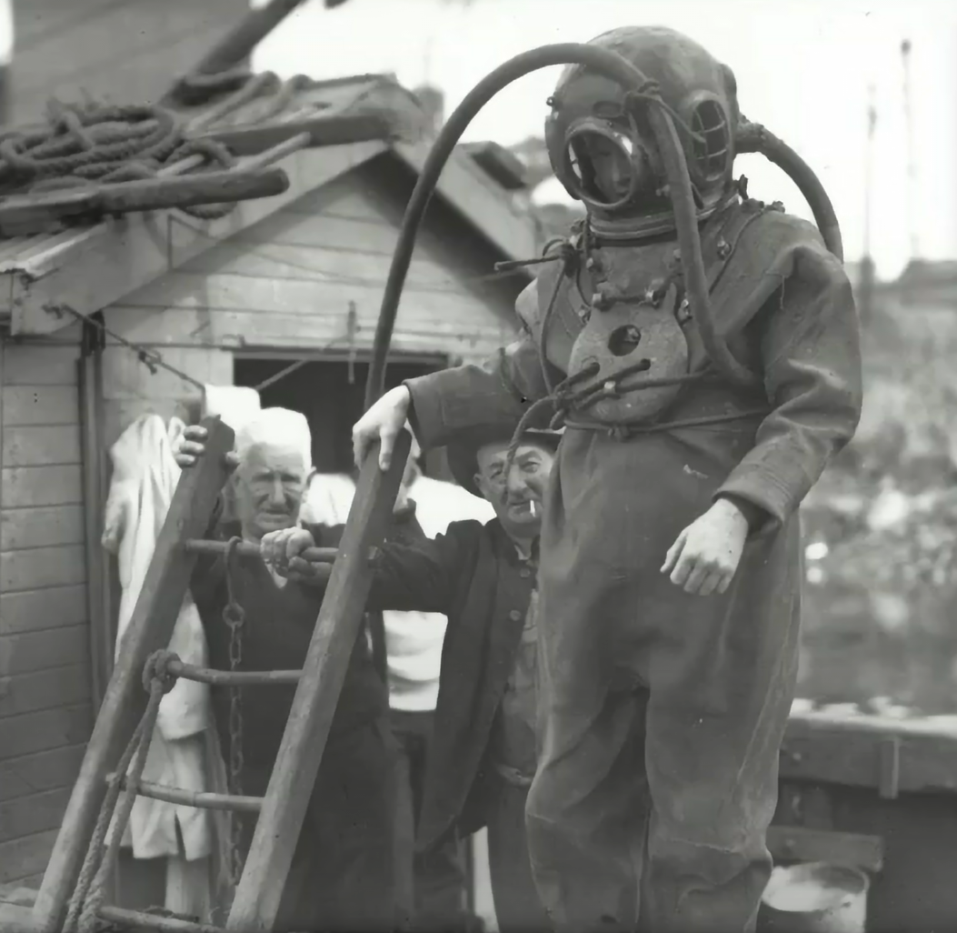 Man in hard hard diving helmet and suit stands at a ladder on a punt. Two men are ready to assist behind him
