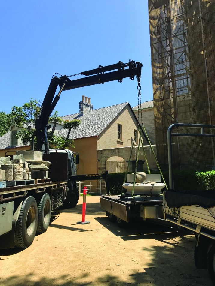 A crane truck lowers stone pieces in front of a scaffolded building