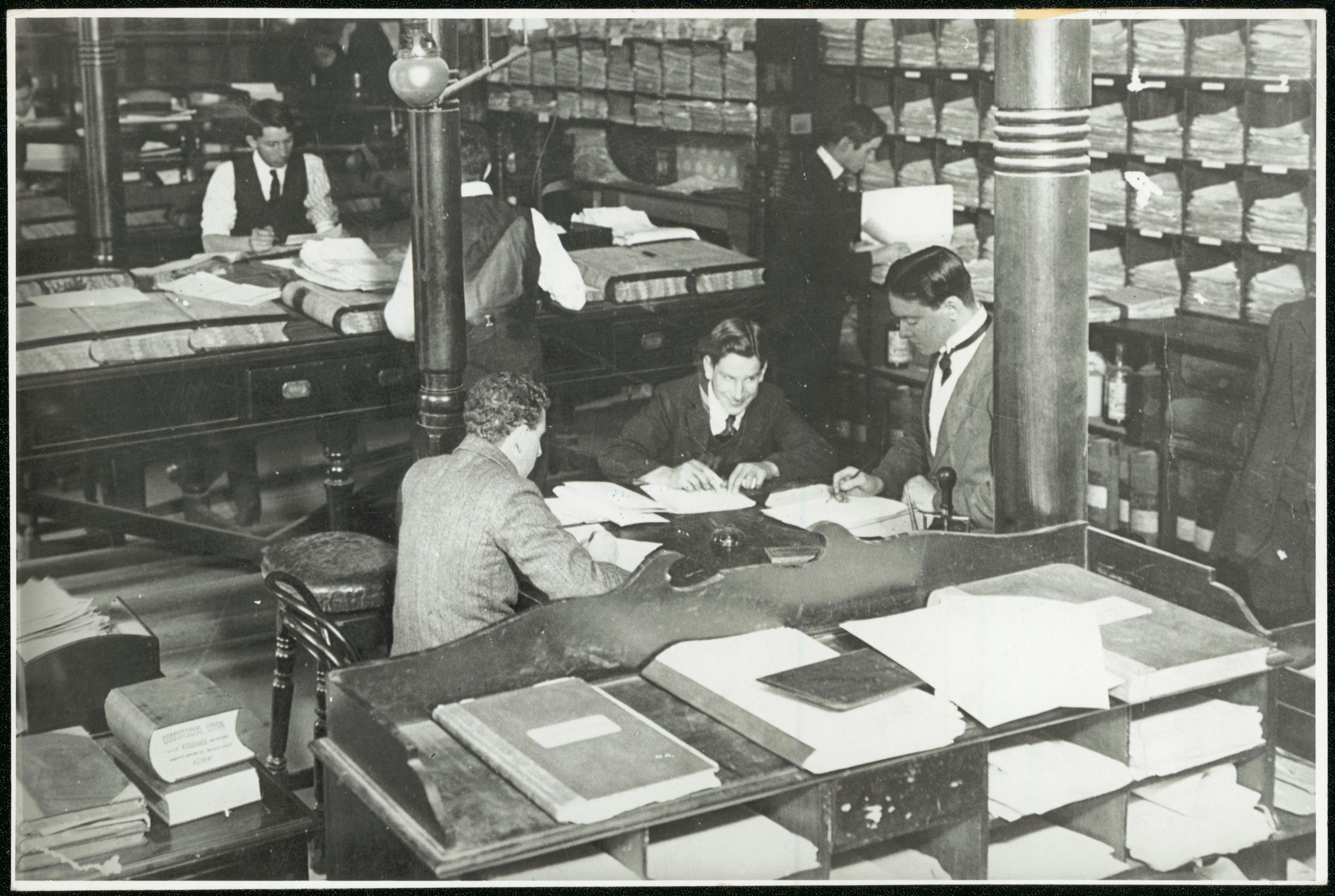 Black and white photo of clerks working at desks in an office, surrounded by large ledgers and pigeon holes and paperwork.