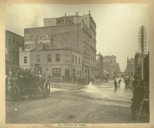 A policeman and spectators look on as the street is washed down with a fire hose