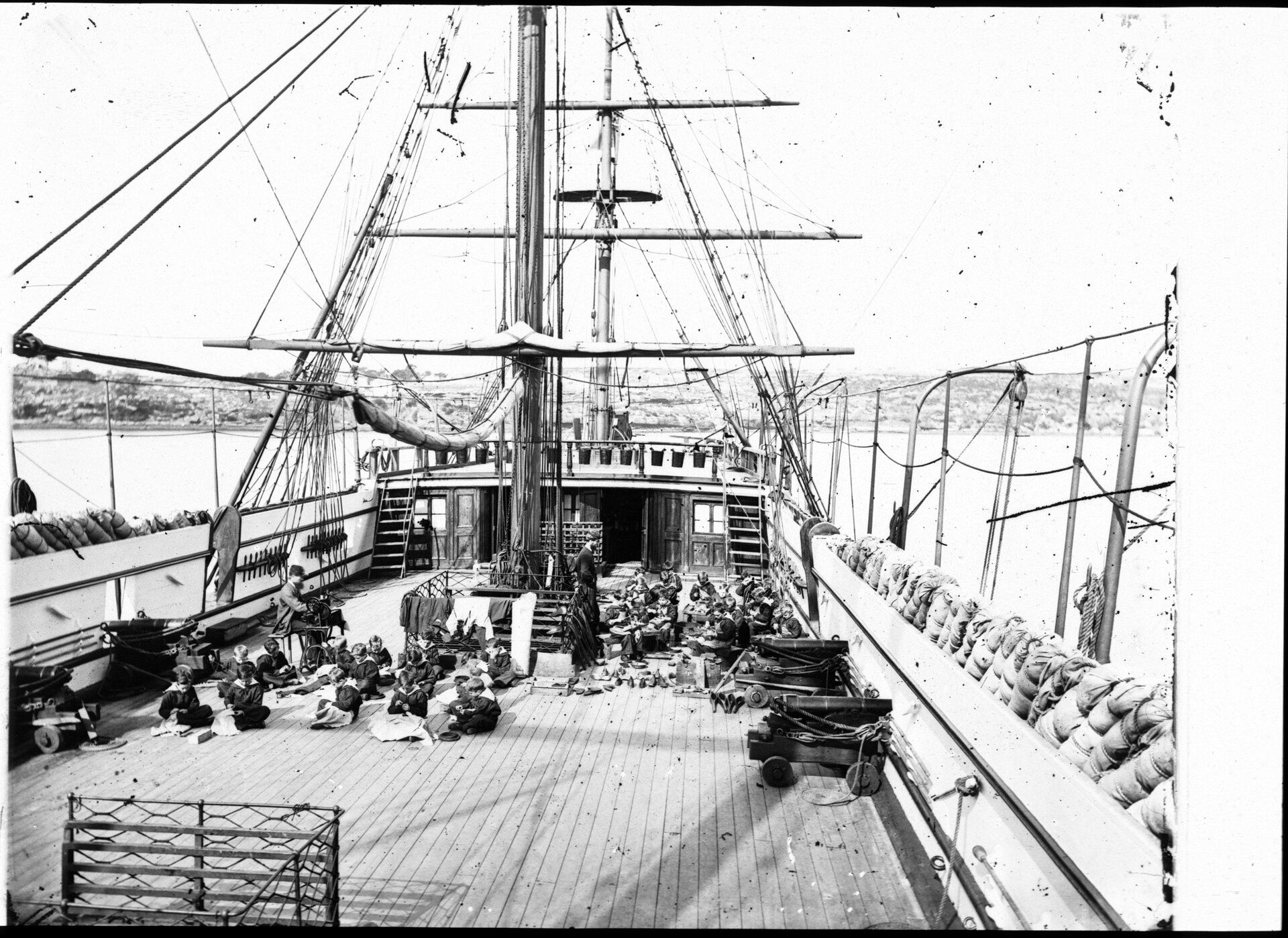 Boys sit cross legged on the deck of a ship with sewing materials while instructors watch on. Cannons line the sides of the deck