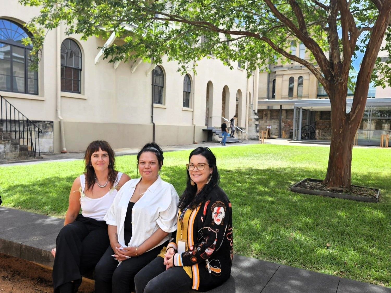 Three women sit on a sandstone fence in front of a building