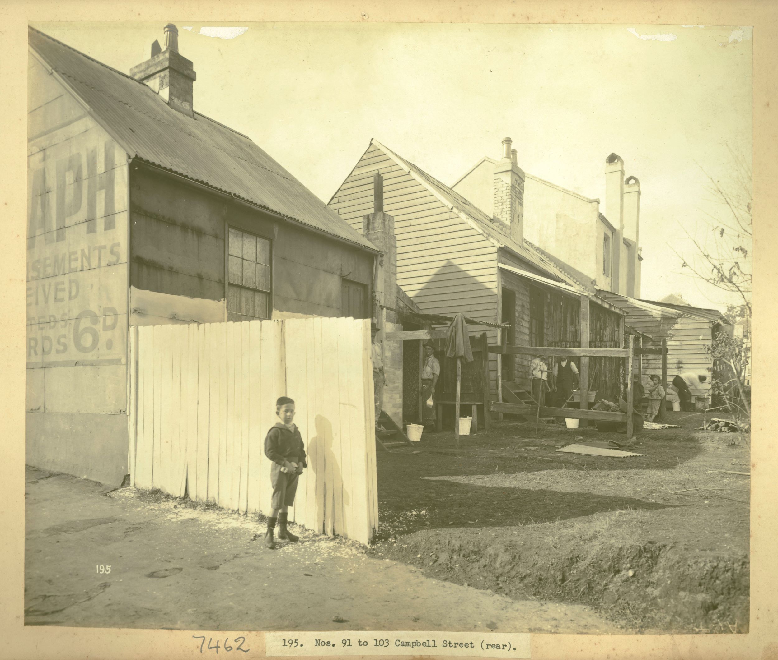 A boy stands in front of a freshly painted white fence and in the yards men stand with cleaning tools and buckets 