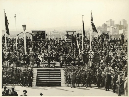 A crowd watches a man speak from a podium that is decorated with the union jack to