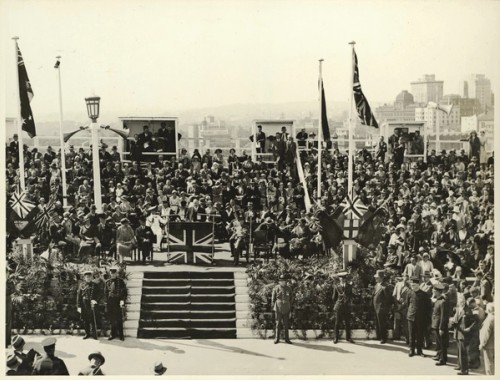 A crowd watches a man speak from a podium that is decorated with the union jack to 