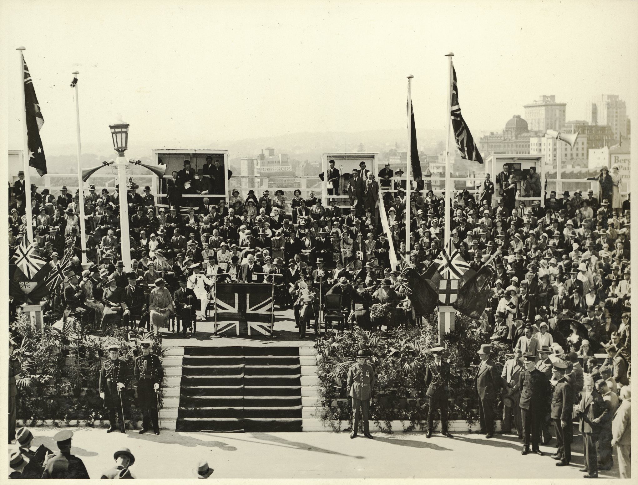 A crowd watches a man speak from a podium that is decorated with the union jack to 