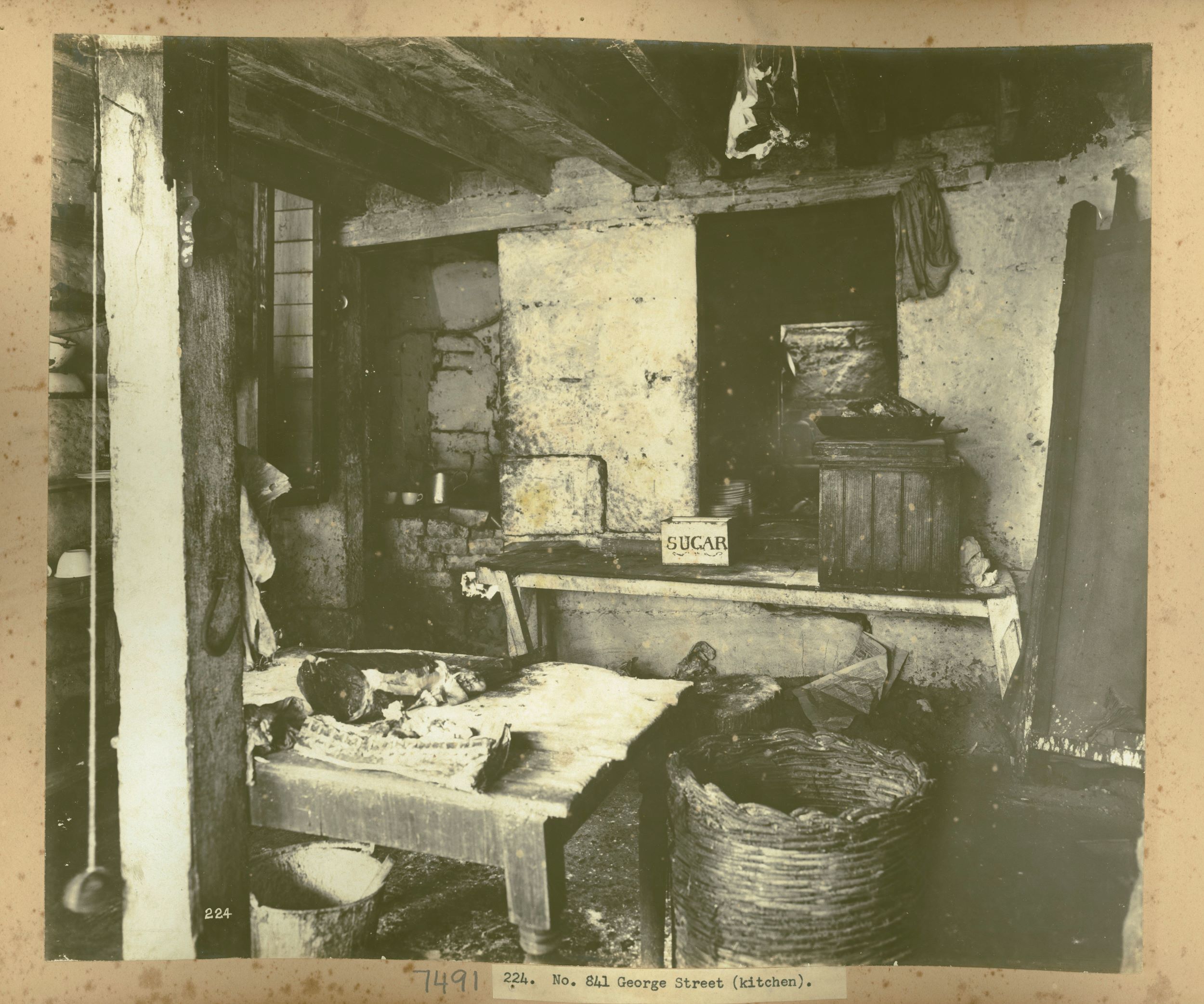 An old kitchen with dirty benches and walls. Cuts of meat sit on a work bench next to a large cane basket 