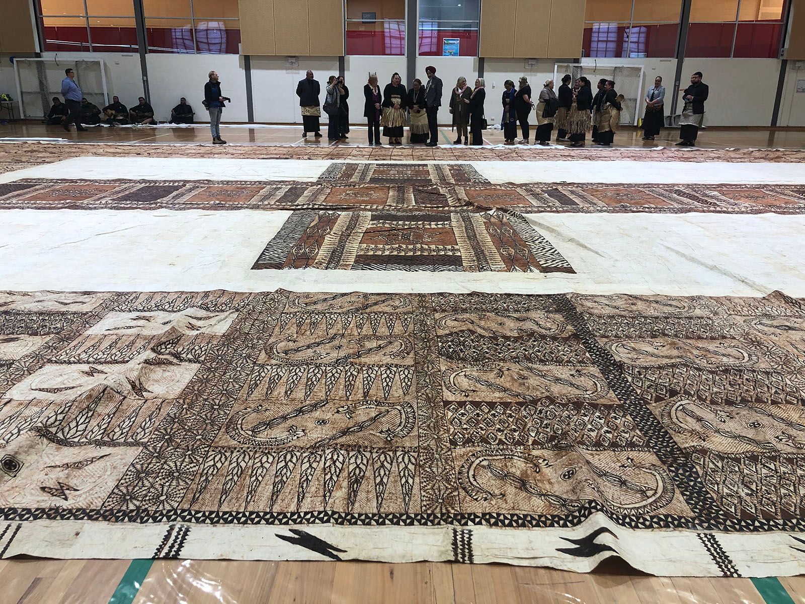 In a large interior space a decorated cloth covers the floor. It is an off-white colour, and the foreground and background borders are decorated in varying [traditional Tongan?] patterns painted in black and shades of brown. At the centre a large white rectangular section is decorated with a cross shape painted in patterns of the same but darker colours. A row of people, several wearing elements of traditional costume wrapped around their waists, stand on the far side of the cloth.