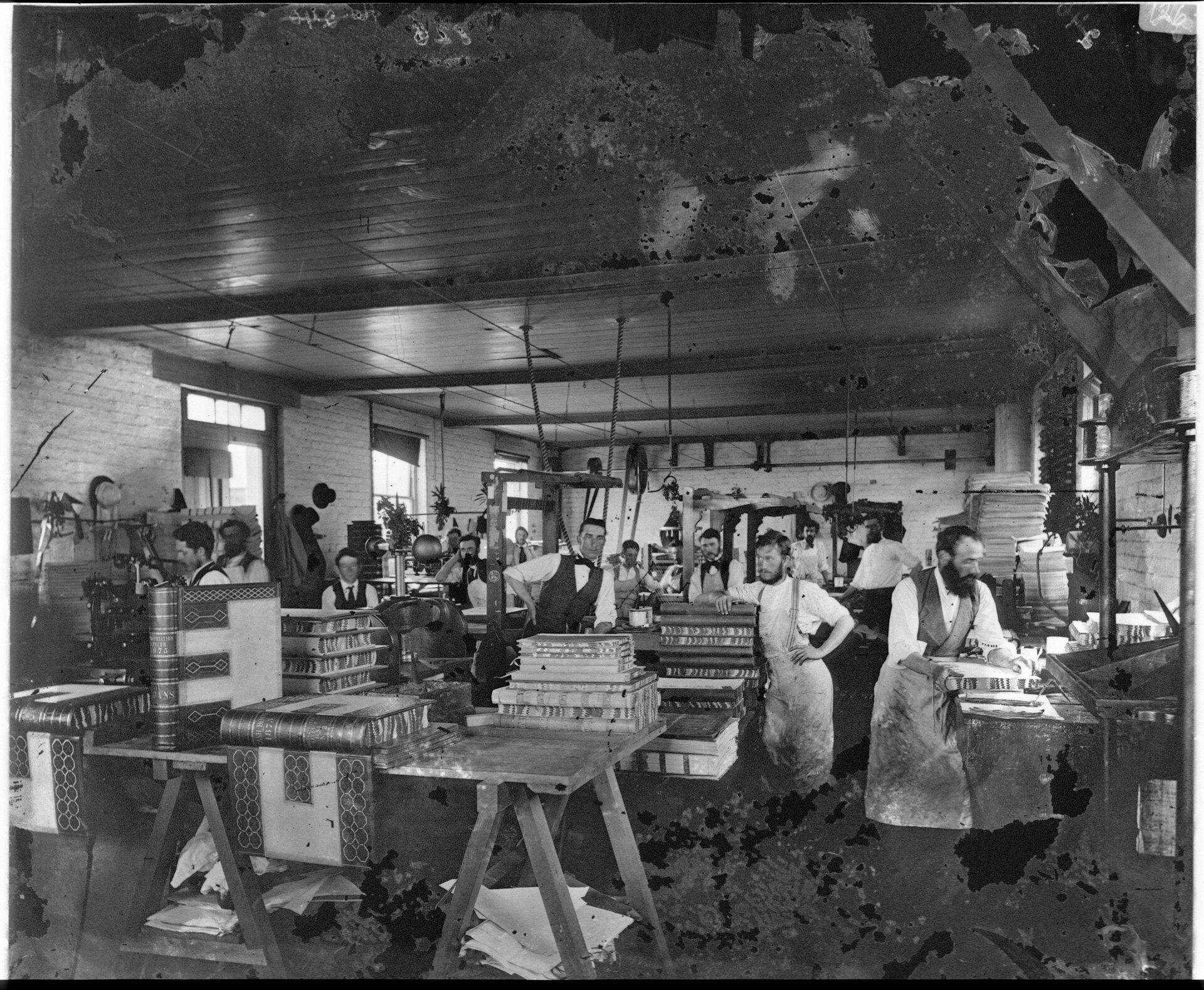 Men in work aprons stand in a workshop surrounded by tables stacked with large books