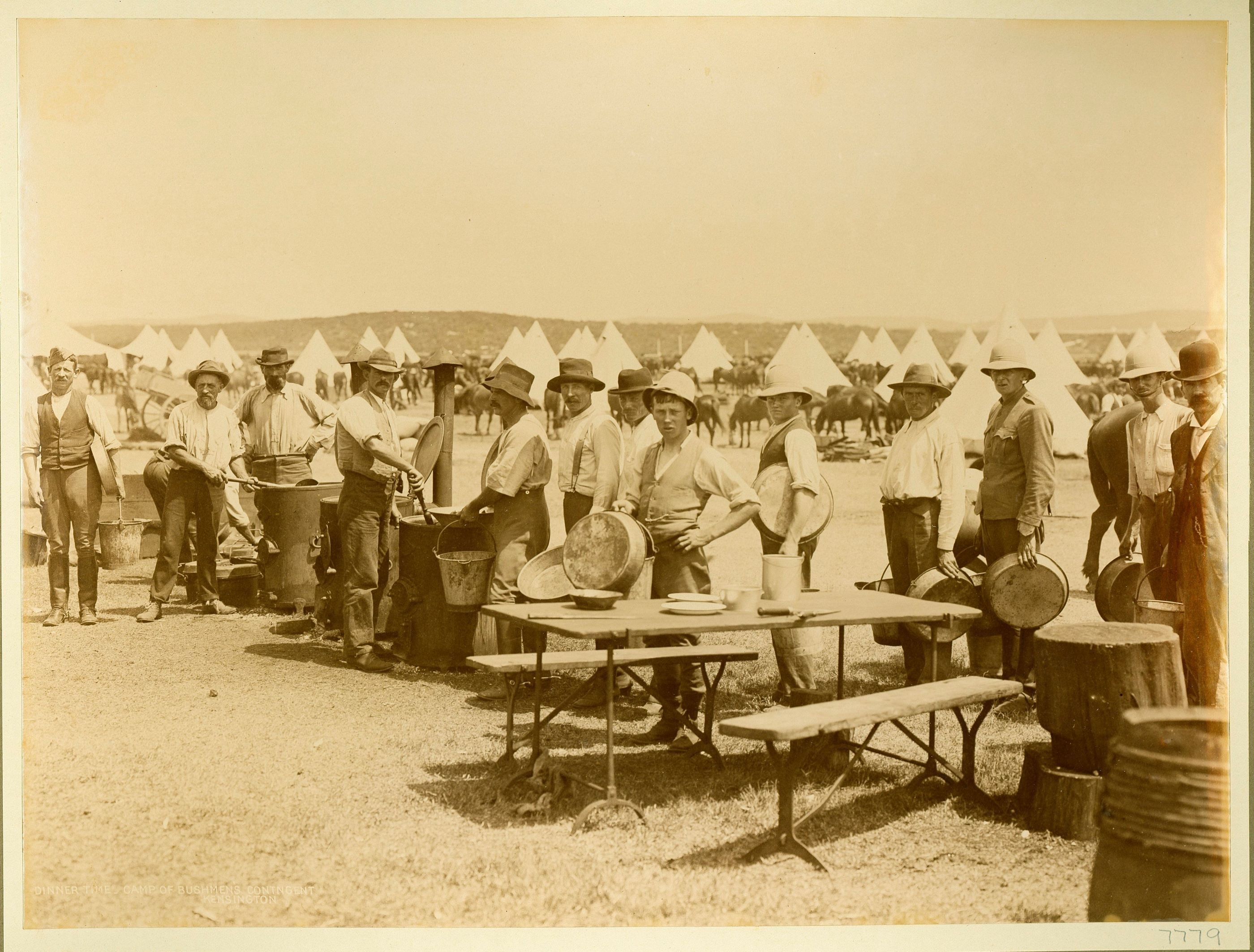 Men stand in a line behind a table and bench seats holding metal bowls 