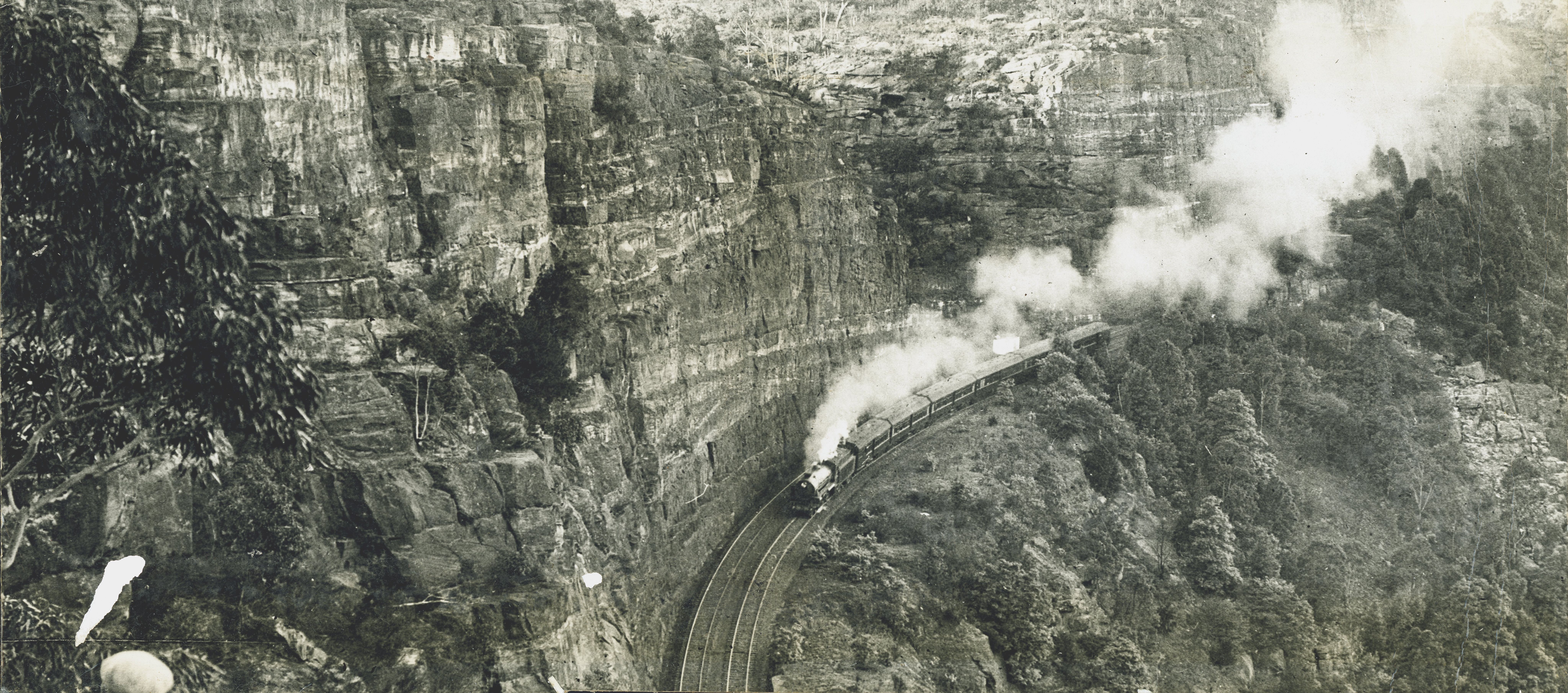 A steam train travels along a railway cutting with a cliff face on one side and a cliff drop on the other