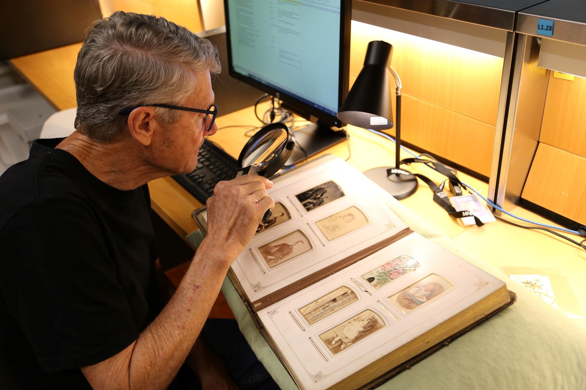 A man sits at a desk and looks at an album of pictures through a magnifying glass