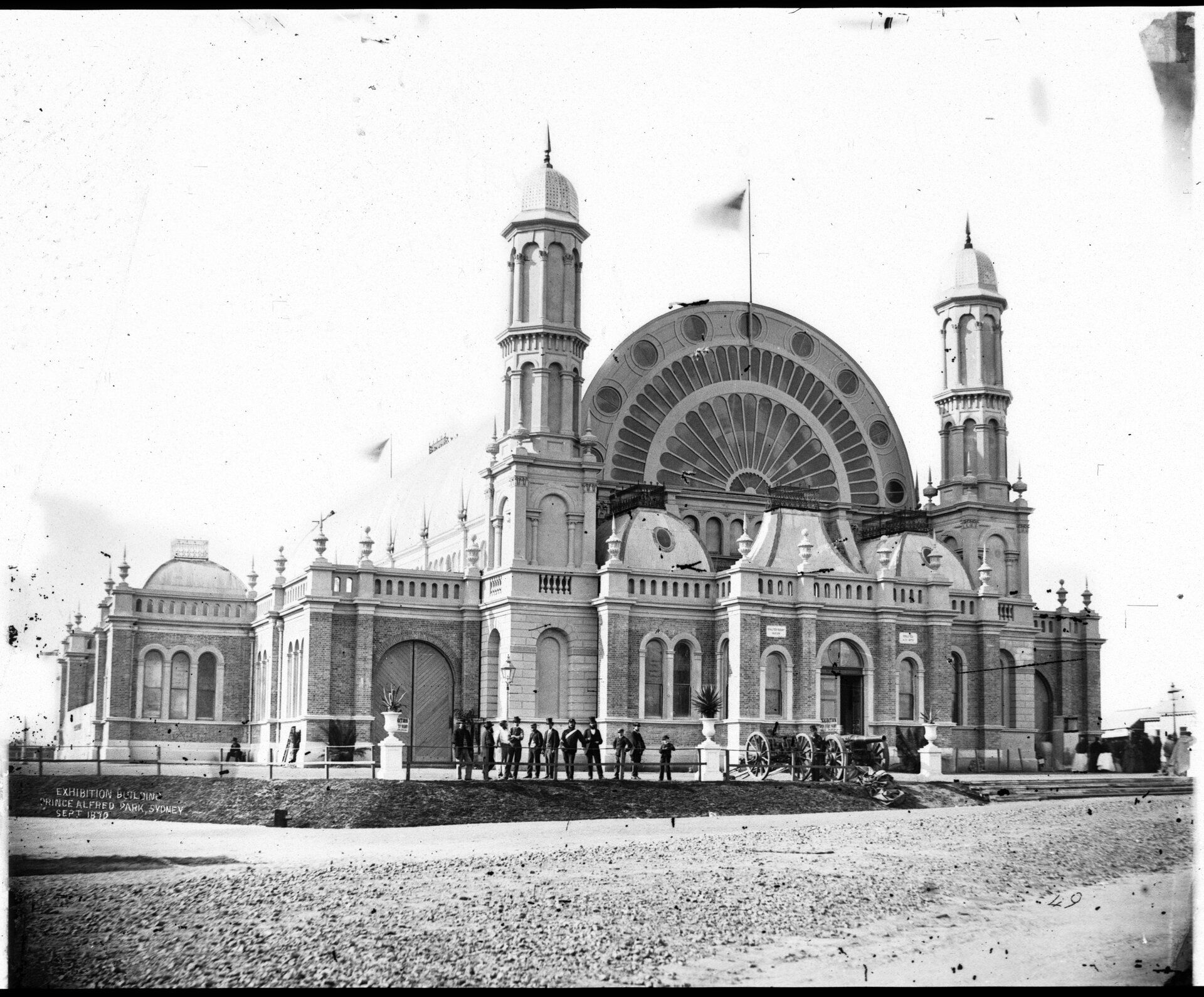 A domed exhibition building with two large towers on either side. A group of people and a horse and cart stand in front