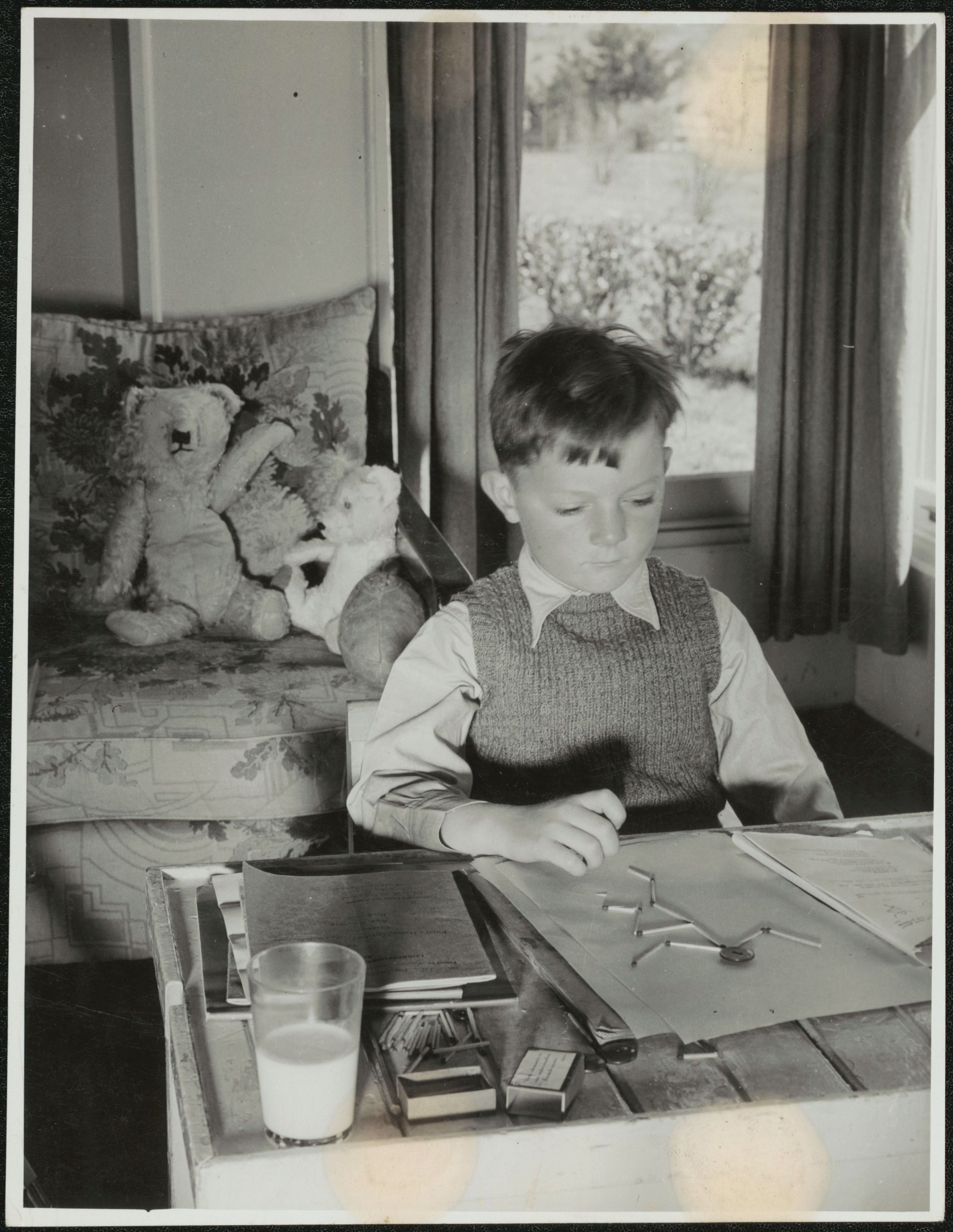 A boy sits at his desk making a match stick man. A glass of milk is on the desk and his teddy bears sit on a chair behind him