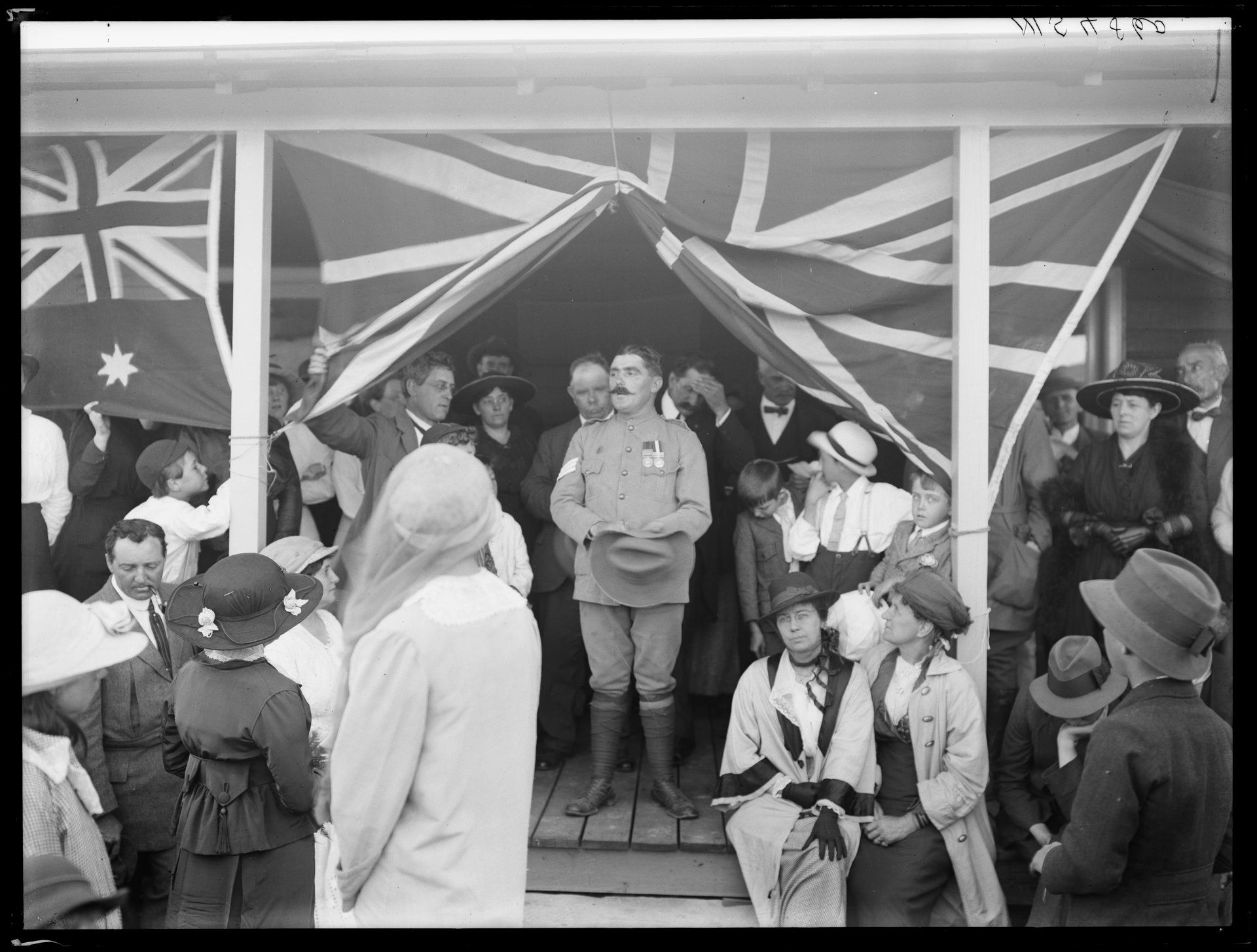 A soldier stands on a stage with the British and Australian flags decorating either side. A crowd is before him and several people stand behind him 