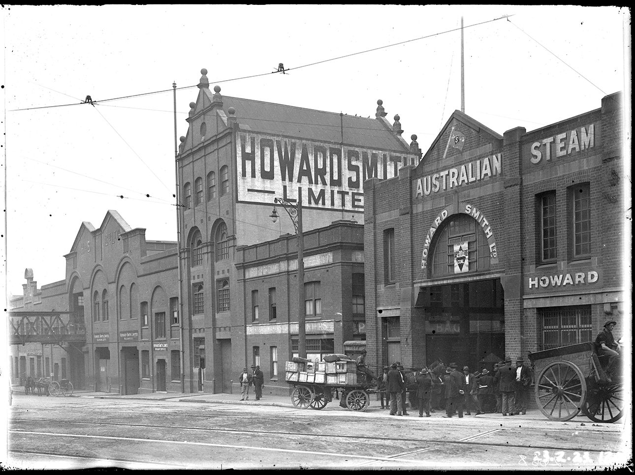 A group of men stand in front of a row of office buildings near a horse and cart loaded with goods