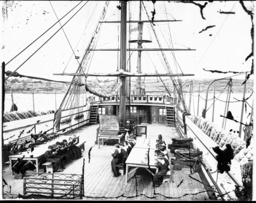 Boys in nautical uniforms sit a long benches on the top deck of a ship and a man at a blackboard stands in the centre near the mast