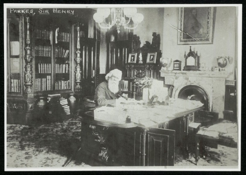 A man sits at a desk in a large office surrounded by bookshelves and a fireplace