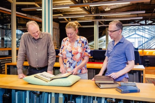Two men and a woman stand at a library shelf looking at items from the collection