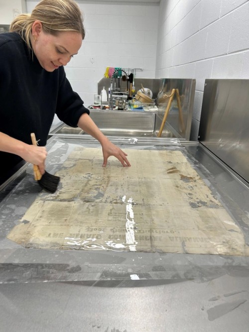  The tissue with applied paste was then placed with the adhesive side down onto the back (verso) of the item. Traditionally, this step may have been completed using a long wooden stick, but here we’ve used a sheet of mylar (polyester) to position the lining sheet on the item. Abby then used a Japanese smoothing brush to ensure the tissue has a good bond to the item.  The item was dried in between blotters and under weight for a few days.