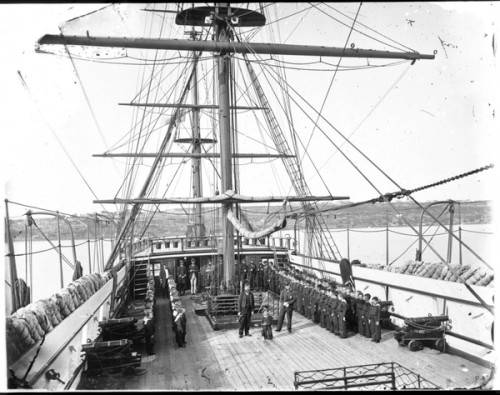 Boys in nautical uniforms stand at attention in straight lines on either side of the top deck of a ship