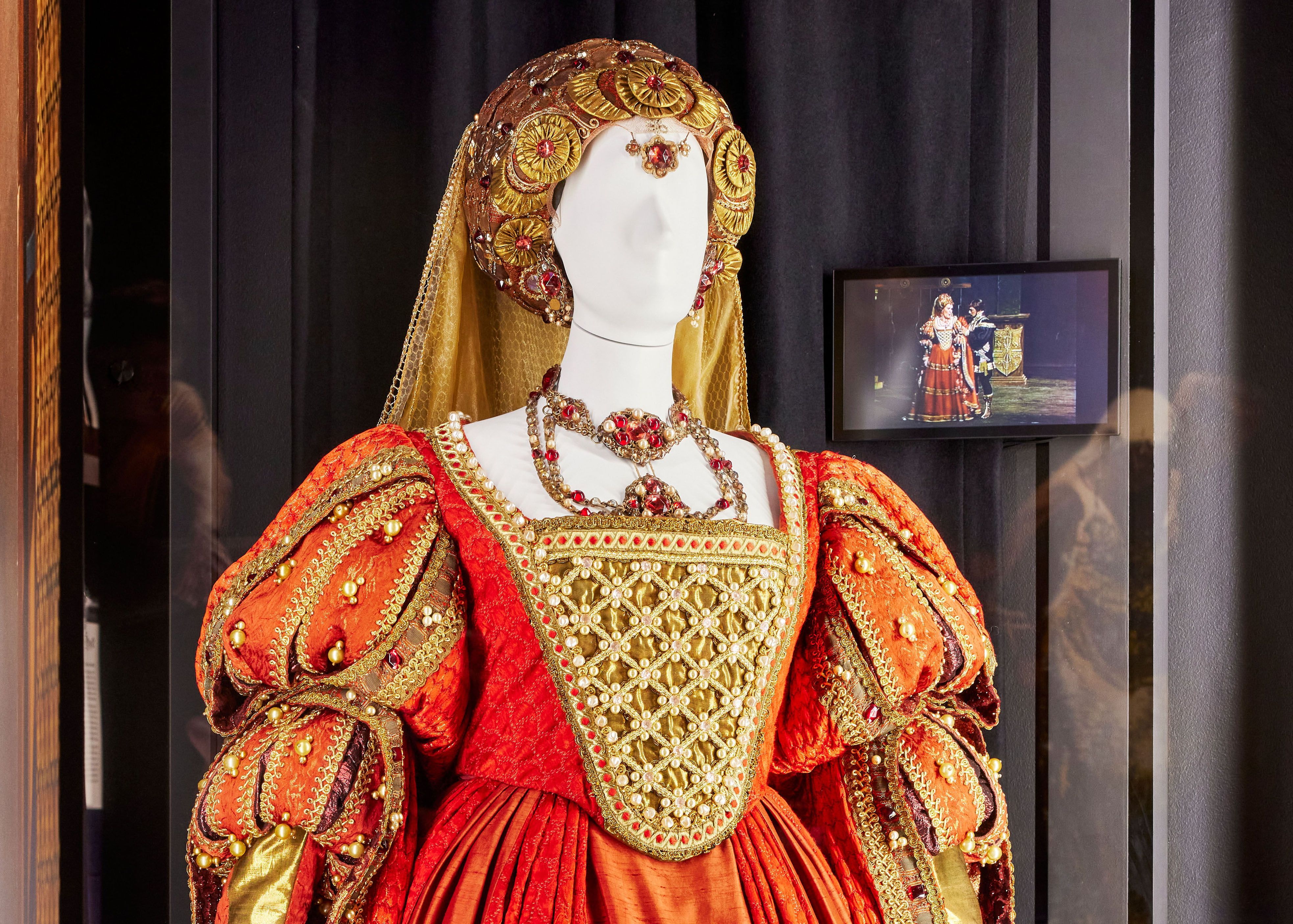 A mannequin dressed in an elaborate Renaissance-style stage costume stands in a large Perspex-fronted display case . On the rear wall a small screen is showing a scene from the opera Lucrezia Borgia where Dame Joan Sutherland is wearing the same costume. 