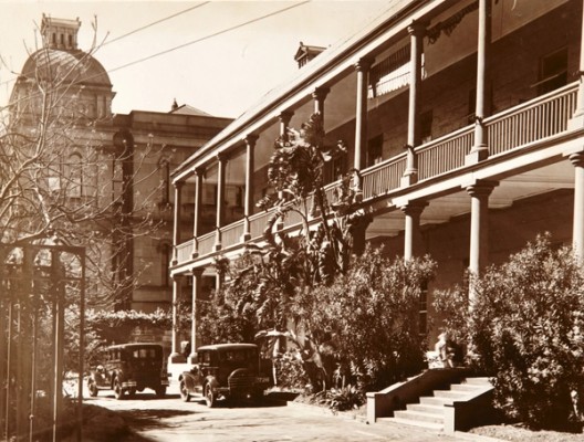 Large 2 storey building with deep verandahs, steps leading to lower verandah and bushes and driveway in the foreground.