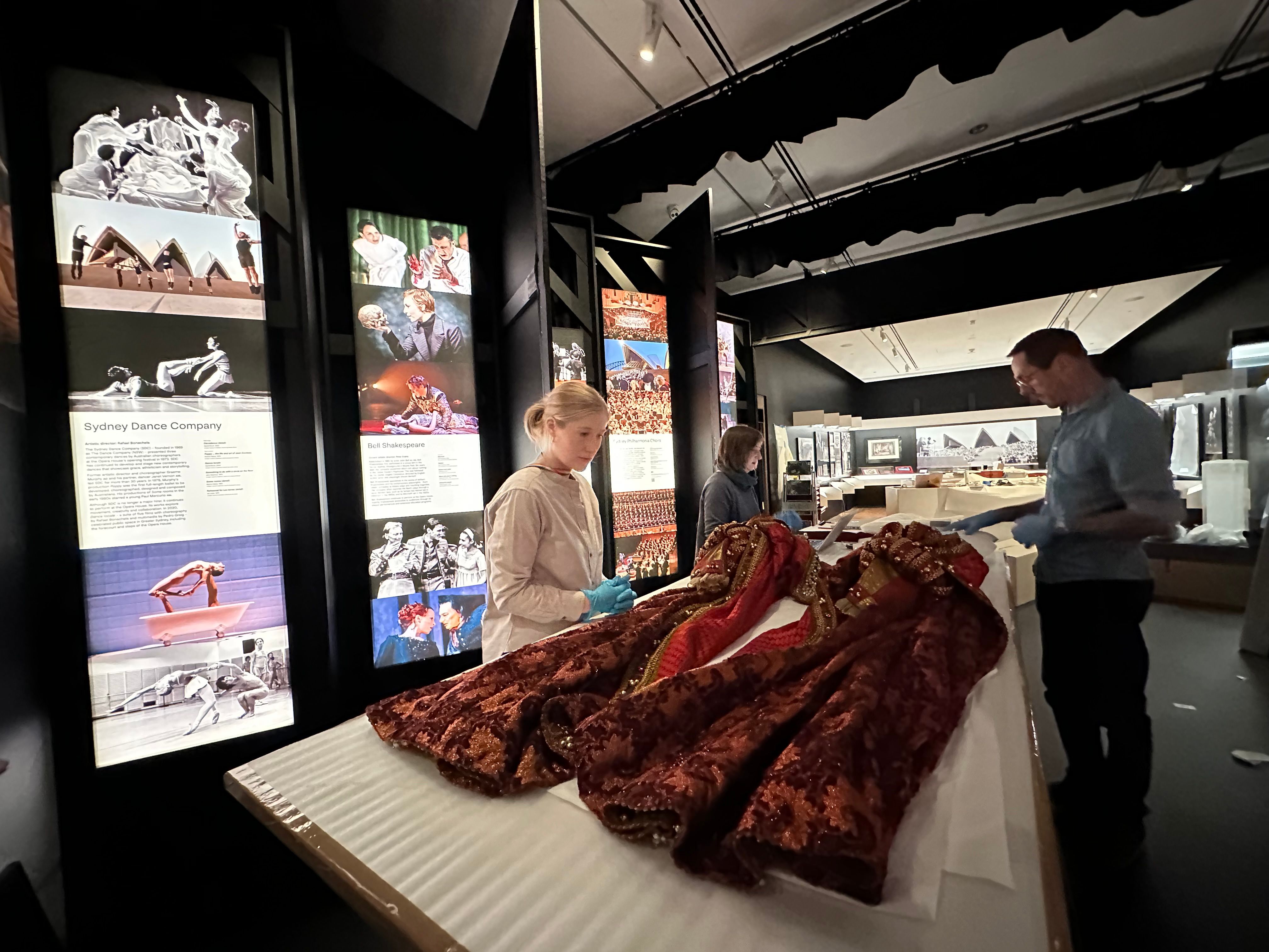 A large, elaborately decorated red dress is stretched out on a large table within an exhibition space, being examined by three people.