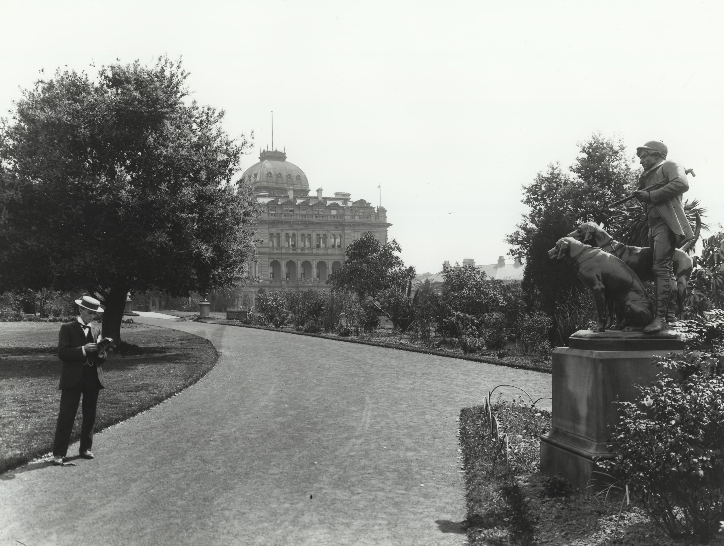 A man in a suit and boater hat stands at the side of the road that to the Colonial Secretary's building