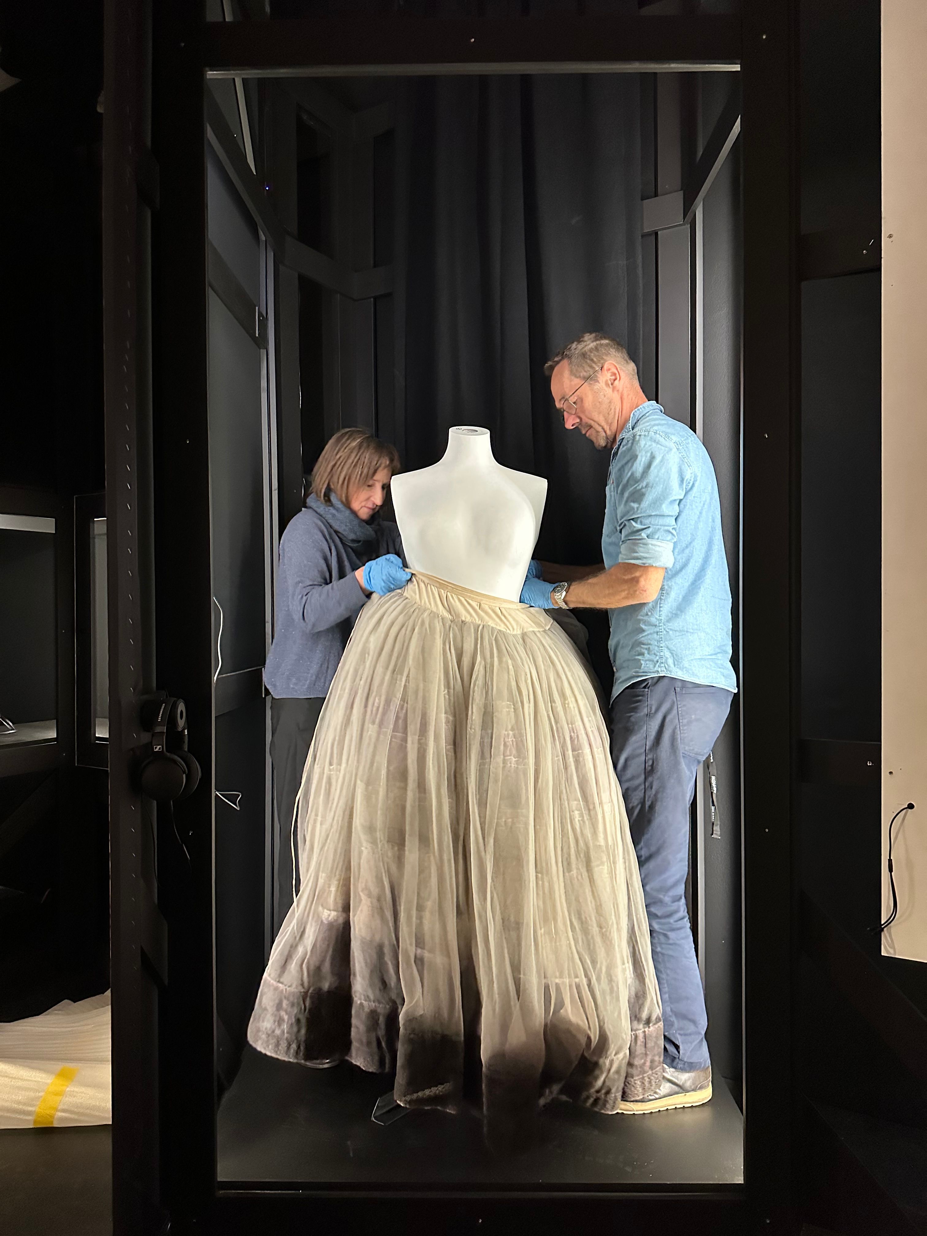A full-length petticoat, coloured off-white with a darker toned band across its base, is being placed on a mannequin by two people, inside a narrow display compartment within an exhibition space.